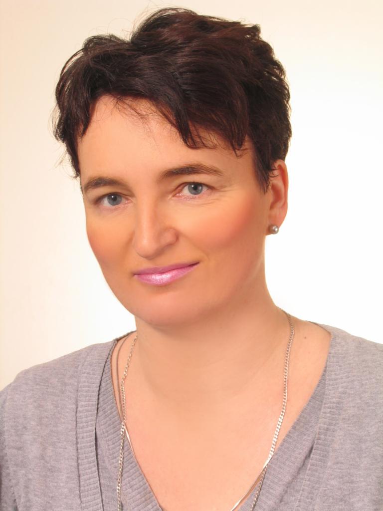 Portret kobiety z krótkimi ciemnymi włosami, ubranej w szary sweter i srebrny łańcuszek, z delikatnym uśmiechem i niebieskimi oczami.