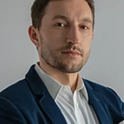 Portret mężczyzny w niebieskiej marynarce i białej koszuli, z chusteczką w kieszonce, na szarym tle.