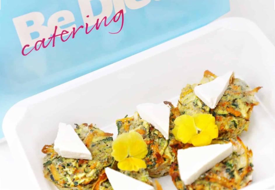 Warzywne muffiny z cukinii i marchewki, udekorowane trójkątem sera i jadalnym kwiatem, ułożone w białym pojemniku z logo firmy cateringowej w tle.