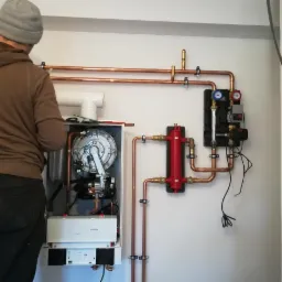 Instalacja nowego pieca grzewczego z miedzianymi rurami i zaworami, widoczny instalator w szarej czapce podczas montażu.