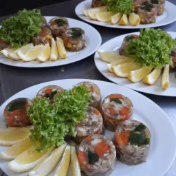 Trzy białe talerze z galaretką wieprzową z warzywami, udekorowaną sałatą i ćwiartkami cytryny, gotowe do serwowania.