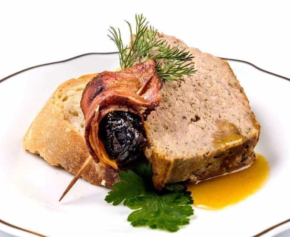 Pasztet z sosem, śliwką w boczku na wykałaczce i kromką chleba, udekorowany koperkiem i pietruszką na białym talerzu z brązową obwódką.