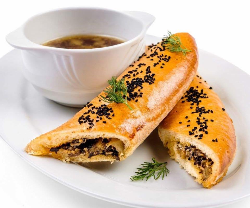 Dwa złociste pierogi z farszem grzybowym posypane czarnym sezamem, udekorowane koperkiem, podane z bulionem w białej miseczce na białym talerzu.