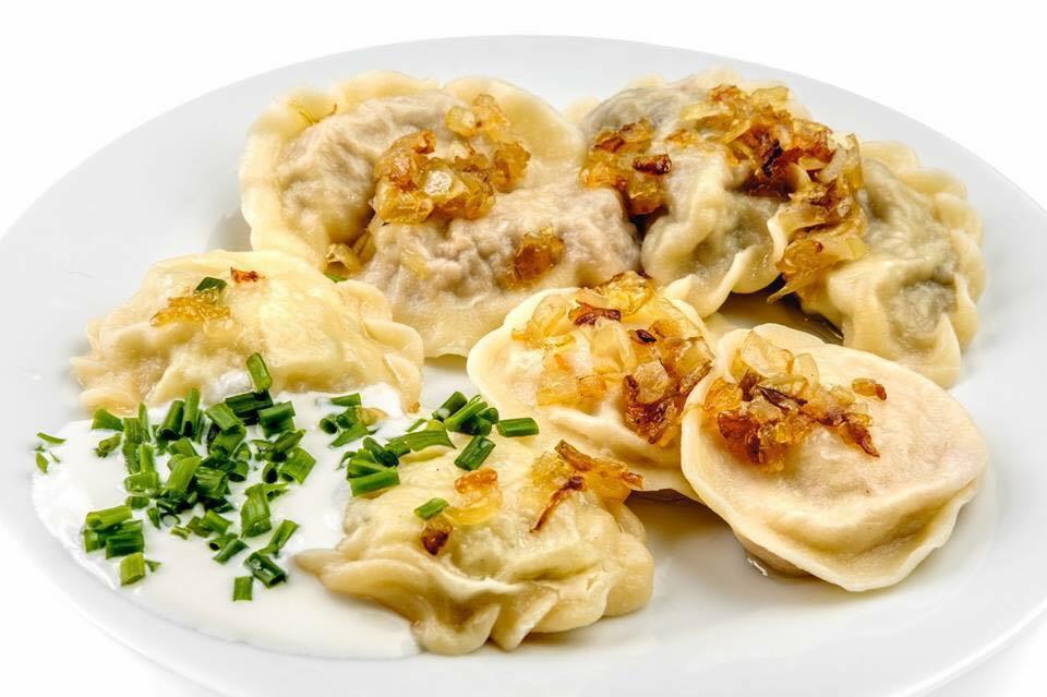 Porcja pierogów z farszem mięsnym i kapustą, polanych podsmażoną cebulką i posypanych świeżym szczypiorkiem, podane na białym talerzu.