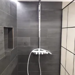 Nowoczesna kabina prysznicowa z deszczownicą i ciemnoszarą okładziną imitującą drewno, z wnęką na kosmetyki i kontrastującymi białymi płytkami.