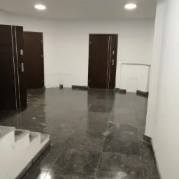 Korytarz w budynku mieszkalnym z szarą, błyszczącą posadzką z płytek, białymi ścianami, dwiema parami ciemnych drzwi i schodami po lewej stronie kadru.