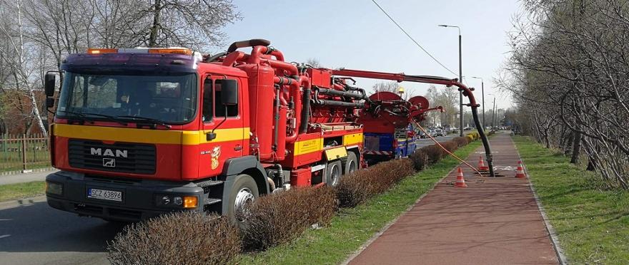 Czerwony samochód specjalistyczny MAN z wysięgnikiem i wężem wpuszczonym do studzienki kanalizacyjnej na chodniku, zabezpieczonej pomarańczowymi pachołkami.