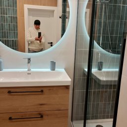 Grzegorz Trąbka - Nowoczesna łazienka z owalnym lustrem podświetlanym LED, drewnianą szafką pod umywalką i kabiną prysznicową z czarnymi profilami. W lustrze odbija się osoba robiąca zdjęcie.