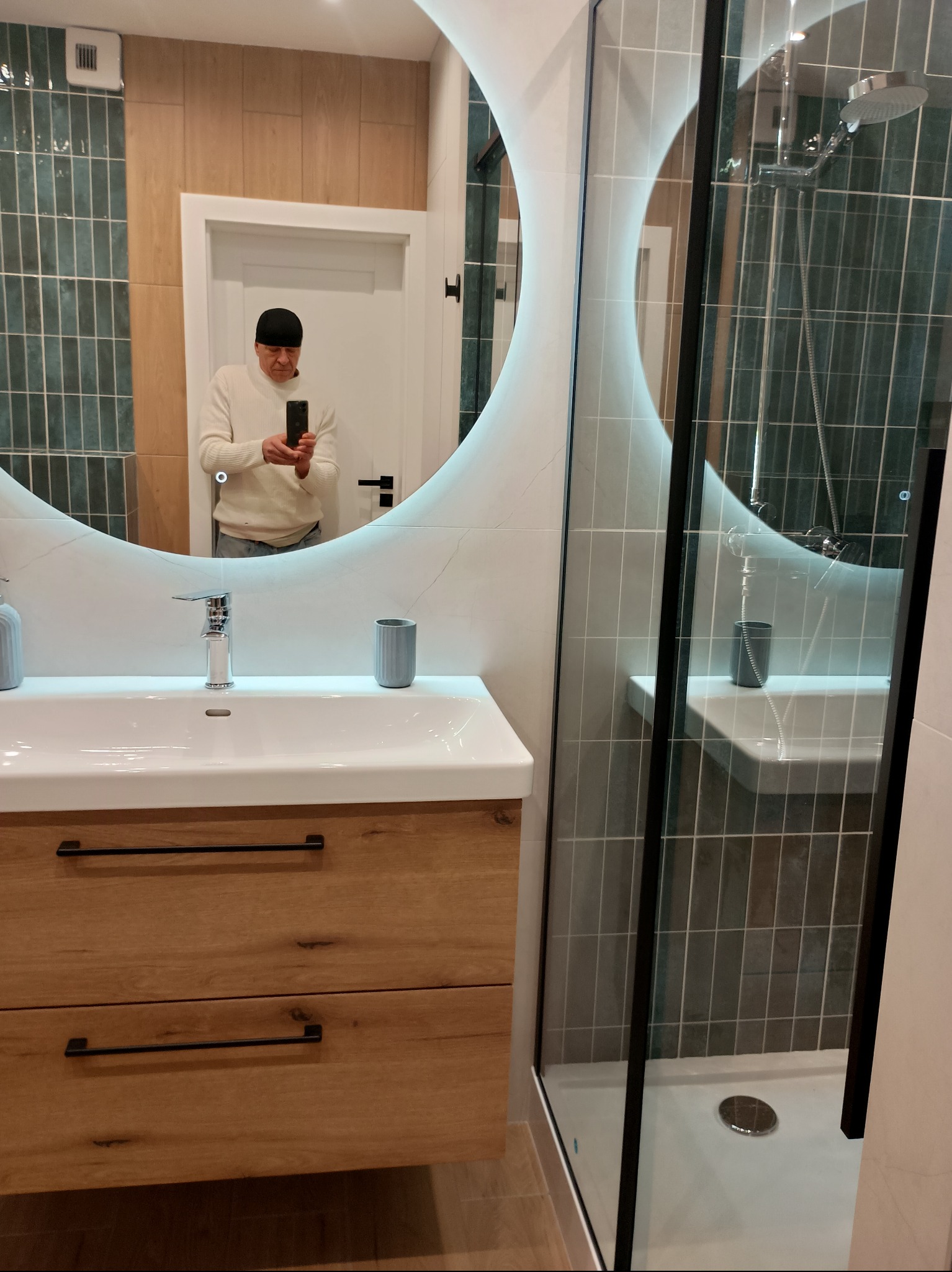 Nowoczesna łazienka z owalnym lustrem podświetlanym LED, drewnianą szafką pod umywalką i kabiną prysznicową z czarnymi profilami. W lustrze odbija się osoba robiąca zdjęcie.