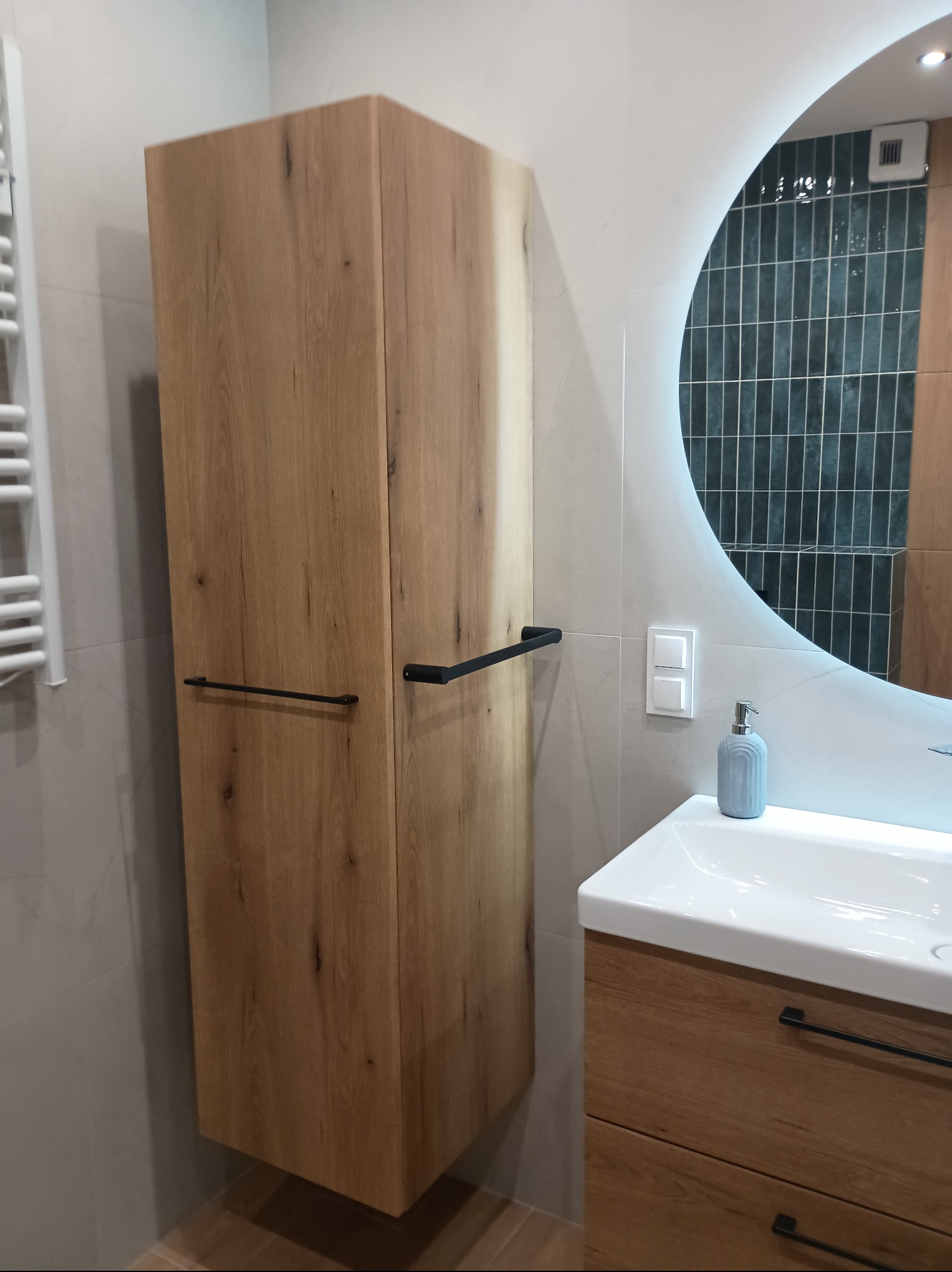 Nowoczesna łazienka z drewnianą szafką wiszącą i umywalką, okrągłe lustro z podświetleniem LED, szare płytki na ścianie i grzejnik drabinkowy.