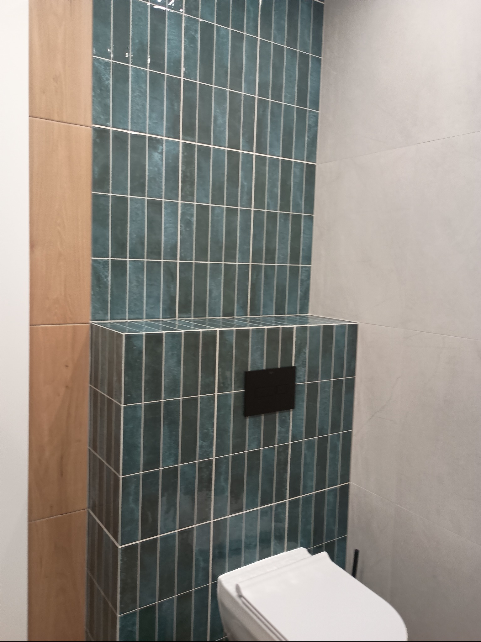 Nowoczesna toaleta z zabudową stelaża pokrytą pionowymi, turkusowymi płytkami. Obok ściana z szarych, wielkoformatowych płyt i fragment drewnopodobnej okładziny.