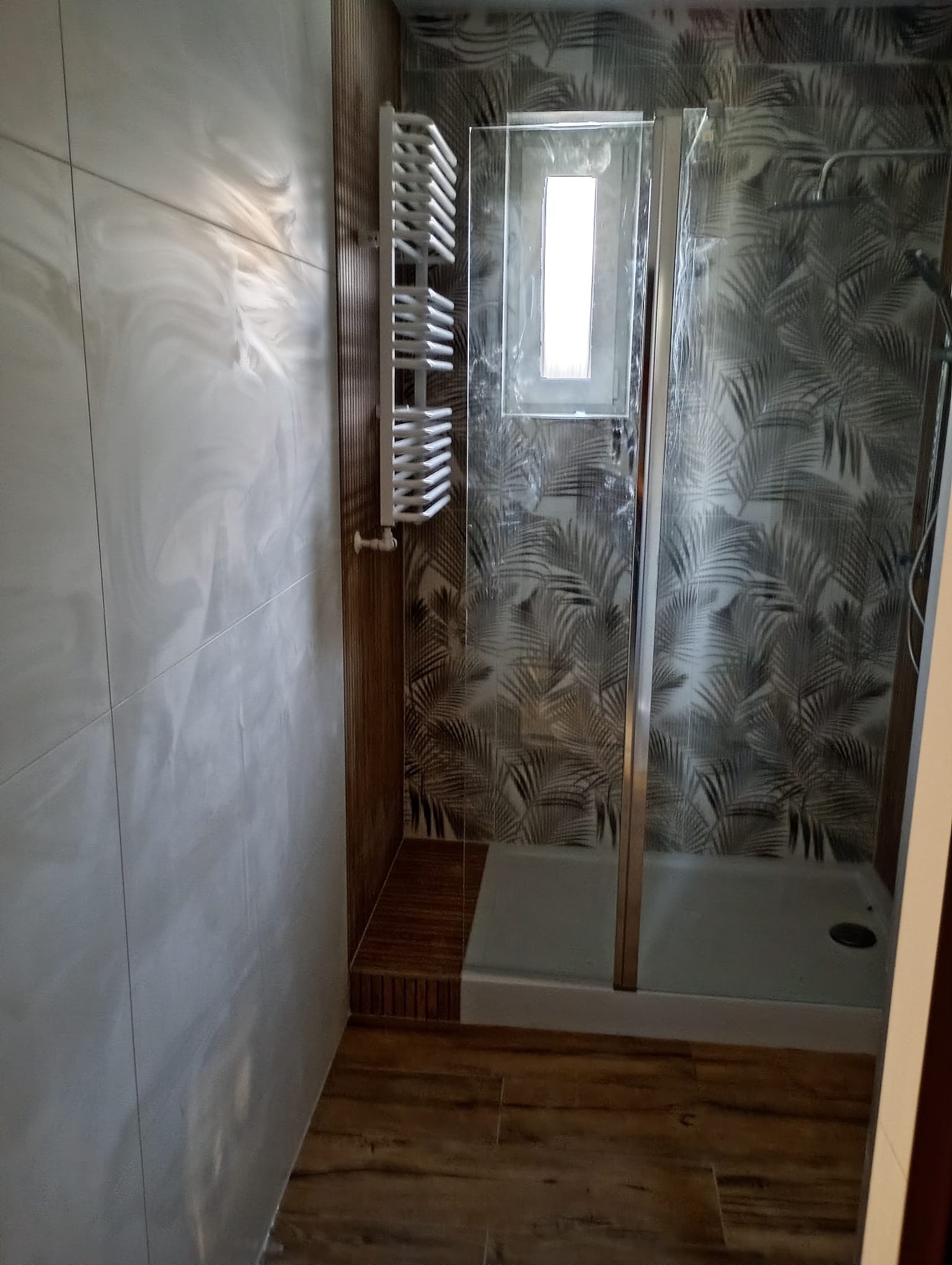 Nowoczesna kabina prysznicowa z motywem liści, grzejnik drabinkowy i płytki imitujące marmur w wykończonej łazience. Widoczne detale wykończenia i oświetlenie.