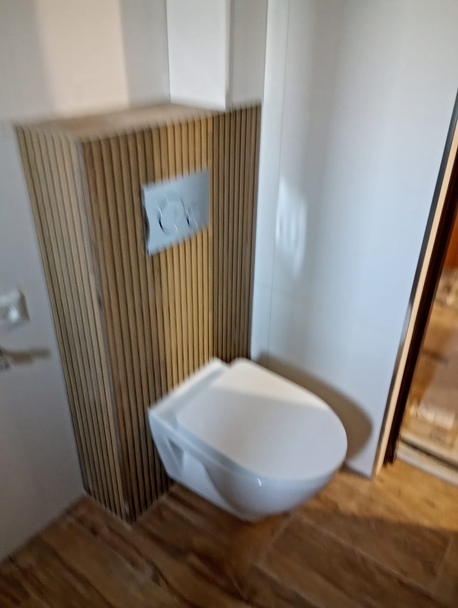 Podwieszana toaleta z zabudową z drewnianych listewek i chromowanym przyciskiem spłukującym, na tle białych płytek i drewnopodobnej podłogi. Ukończona aranżacja.