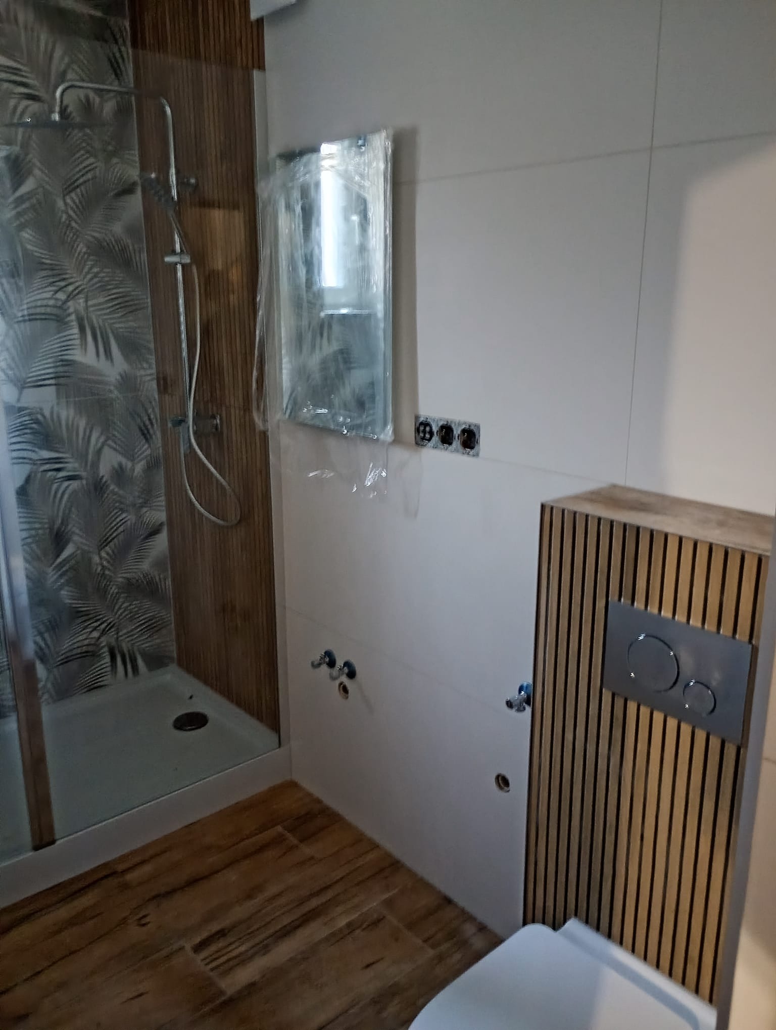 Nowoczesna łazienka z kabiną prysznicową z motywem liści, drewnopodobnymi płytkami na podłodze i obudową toalety w pionowe lamele. Lustro zabezpieczone folią.