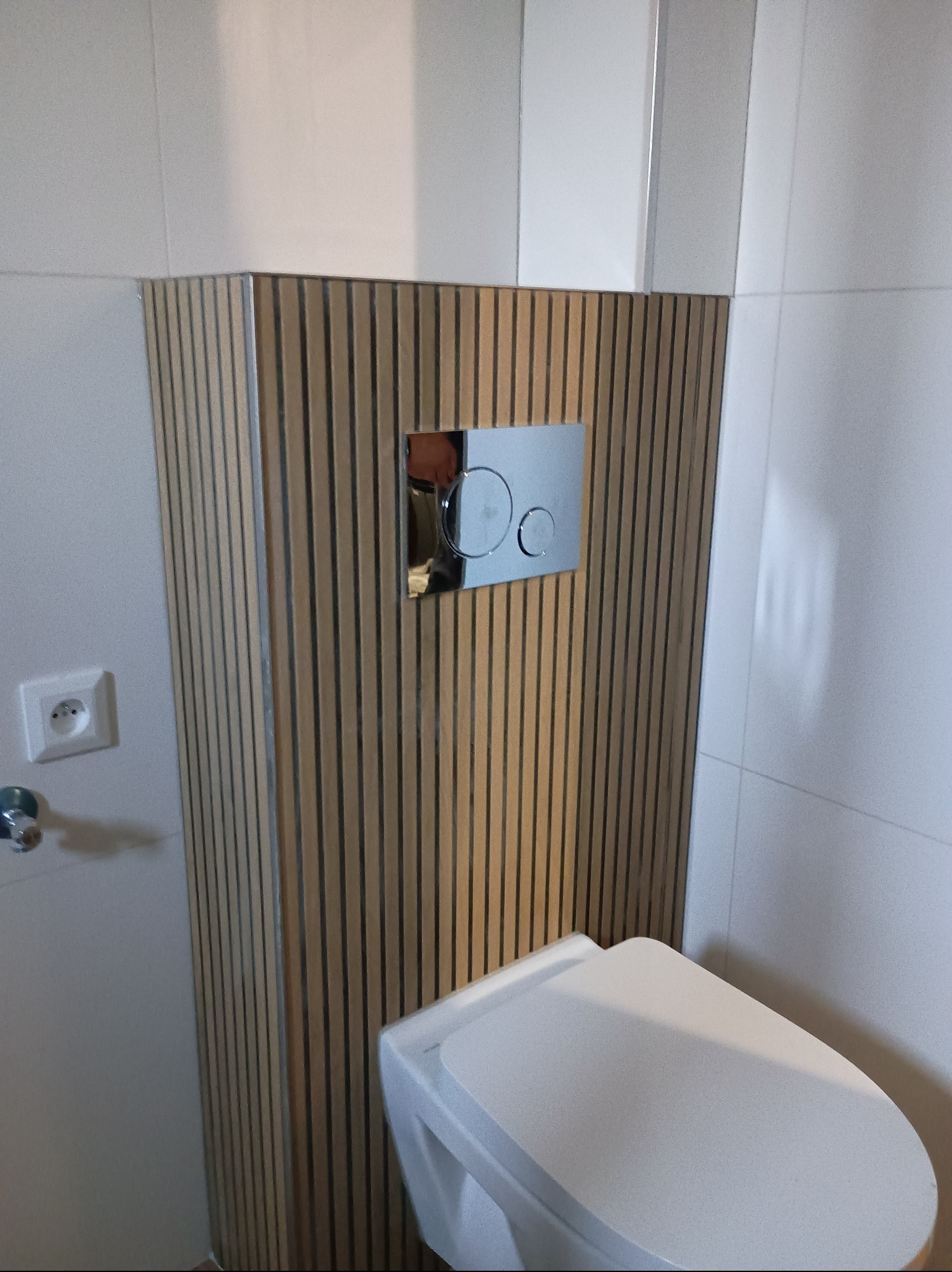 Nowoczesna toaleta z zabudową z drewnianych lameli i chromowanym przyciskiem spłukującym, na tle białych płytek. Widoczny fragment gniazdka elektrycznego.