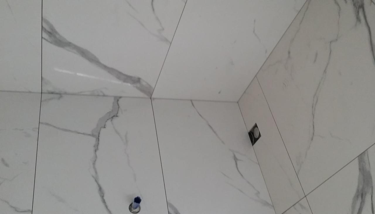 Ściana i podłoga wyłożone białymi płytkami imitującymi marmur z szarymi żyłami, widoczne otwory na instalacje.