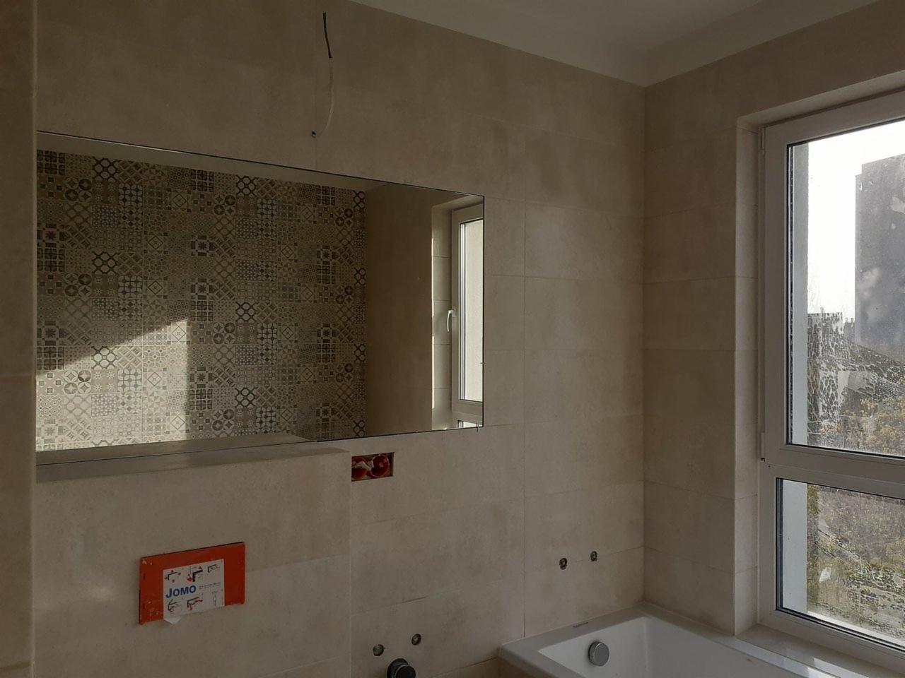 Jasna łazienka w trakcie wykańczania: beżowe płytki na ścianach, lustro odbijające wzorzystą ścianę, okno z widokiem na miasto, widoczna instalacja podtynkowa JOMO.