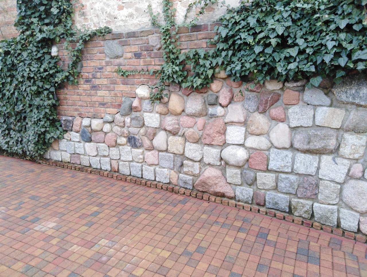 Kamienny mur z elementami cegły, porośnięty bluszczem, z kostką brukową w odcieniach czerwieni i brązu u podstawy.