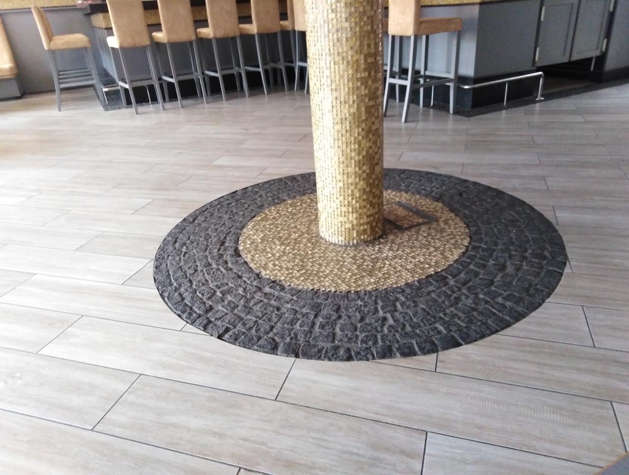Wykończenie podłogi wokół kolumny: jasne, podłużne płytki ceramiczne, okrągły wzór z ciemnych, nieregularnych kamieni i mozaiki wokół kolumny obłożonej jasną mozaiką. Widoczne krzesła barowe w tle.