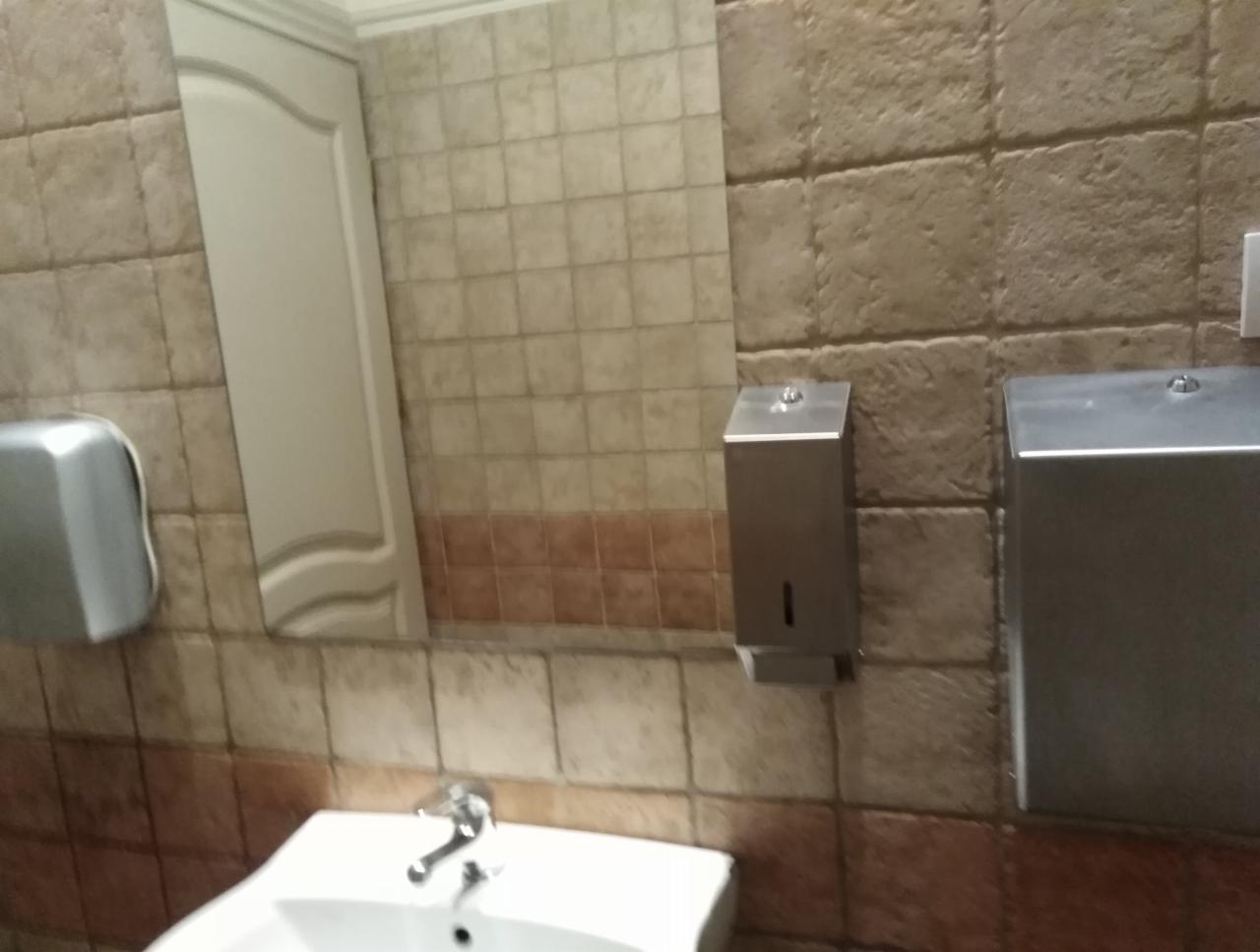 Umywalka z baterią, lustro z białą ramą, suszarka do rąk i dozownik mydła na ścianie wyłożonej beżowo-brązowymi płytkami o nieregularnej fakturze.