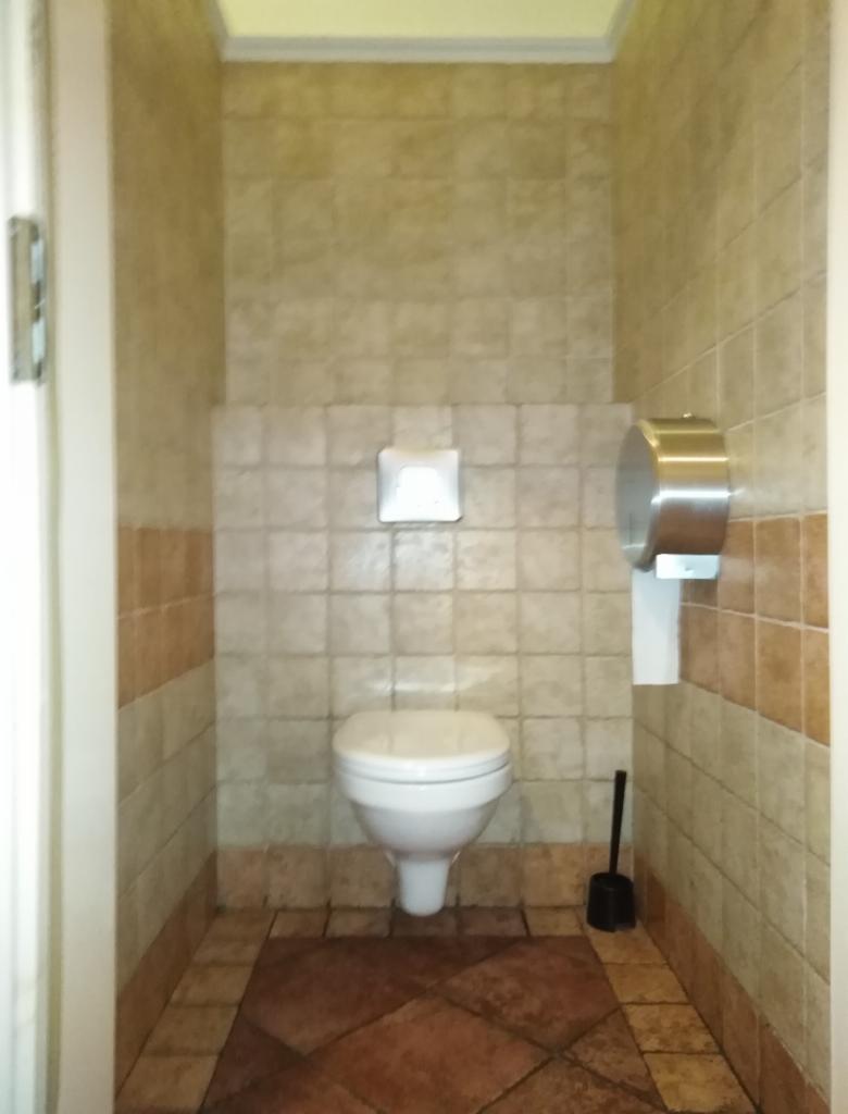 Niewielka toaleta wykończona beżowymi i brązowymi płytkami, z widocznym sedesem, podajnikiem na papier toaletowy i szczotką do WC.