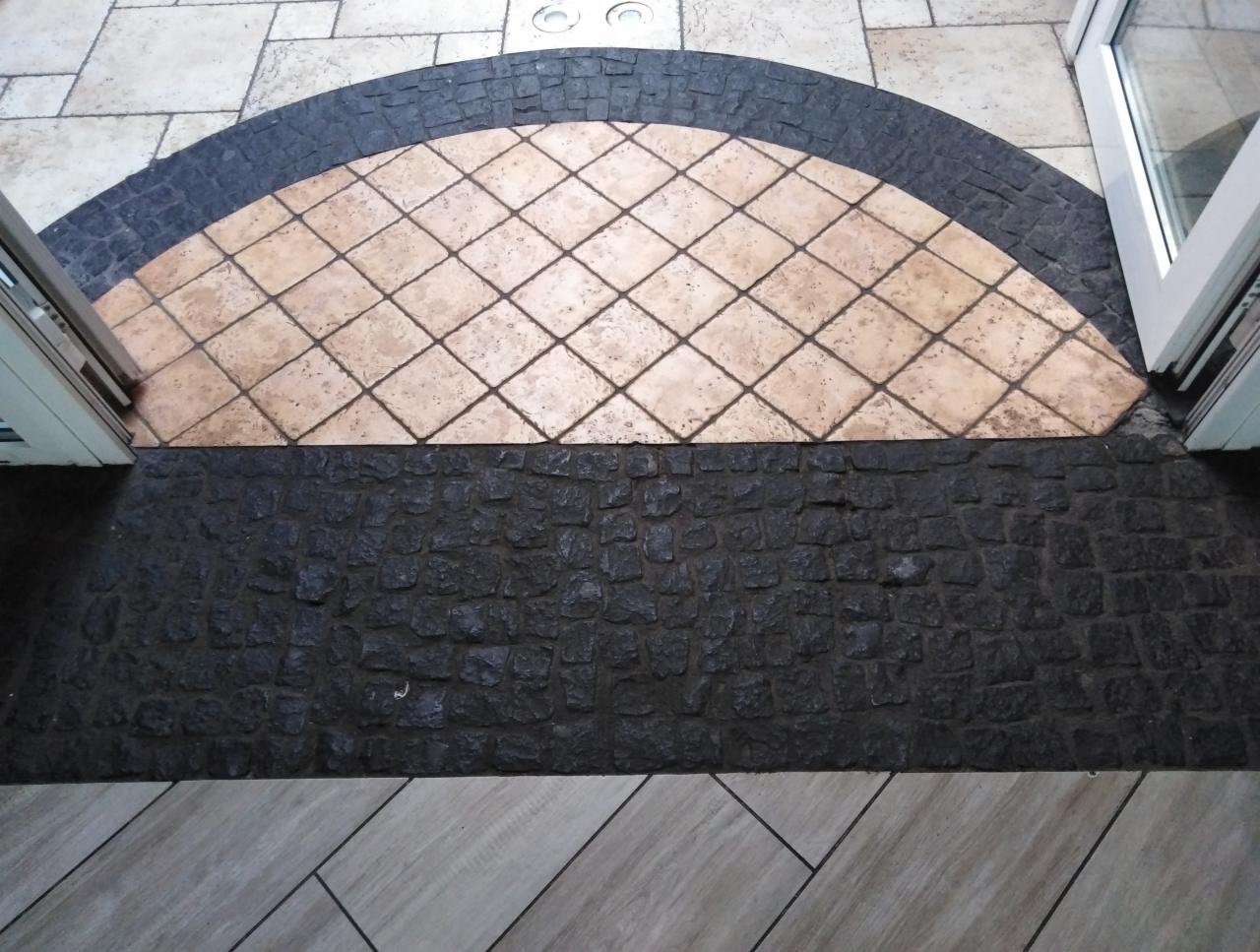 Podłoga z mozaiką z beżowych płytek ułożonych w romby, obramowana ciemną kostką brukową w kształcie półkola, widoczna z perspektywy góry przy otwartych drzwiach.