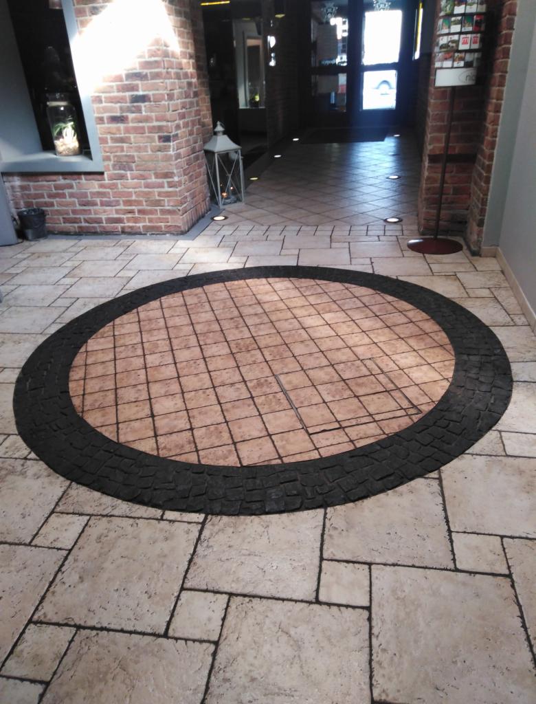 Posadzka z dekoracyjnym okrągłym wzorem z płytek w odcieniach beżu i brązu, otoczona ciemną obwódką z kostki brukowej, widok na korytarz z oświetleniem podłogowym i ceglaną ścianą.