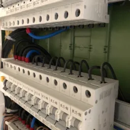 Rozdzielnica elektryczna z zainstalowanymi bezpiecznikami EATON, widoczne przewody w peszlach, kolorowe oznaczenia na końcówkach przewodów, zielona ściana w tle.
