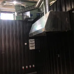 Zamontowany system wentylacji mechanicznej z metalowymi rurami wentylacyjnymi i okapem w pomieszczeniu z panelami ściennymi, widoczne gniazda elektryczne i skrzynka bezpiecznikowa.