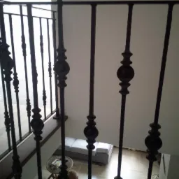 Kute, czarne, zdobione pręty balustrady schodowej, widok z góry na dół, fragment salonu w tle.