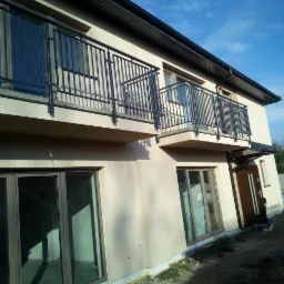 Dwupiętrowy dom z balkonami zabezpieczonymi czarnymi, metalowymi balustradami o prostym wzorze, widok z dołu na elewację i błękitne niebo.