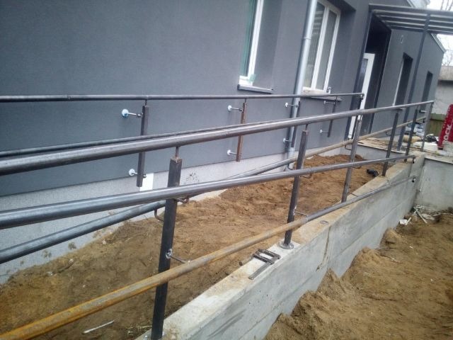 Metalowa balustrada zewnętrzna, surowy montaż przy szarym budynku. Widoczne spawy i elementy mocujące. Ujęcie z dołu, podkreślające konstrukcję.