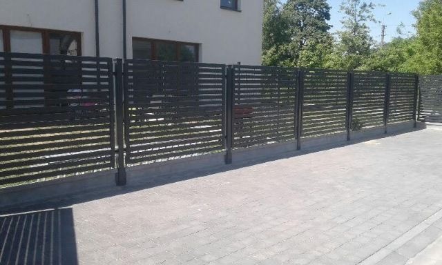 Nowoczesne ogrodzenie posesji z poziomych, metalowych profili w kolorze grafitowym, na tle domu jednorodzinnego i kostki brukowej.