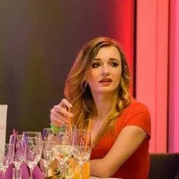 Kobieta w czerwonej bluzce siedzi przy stole z kieliszkami, mieszając drinka słomką. W tle czerwone i różowe oświetlenie.