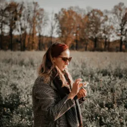 Kobieta w okularach przeciwsłonecznych, w kratowanym płaszczu i szaliku, trzymająca w dłoniach bukiecik drobnych, białych kwiatów na łące w jesiennej scenerii.