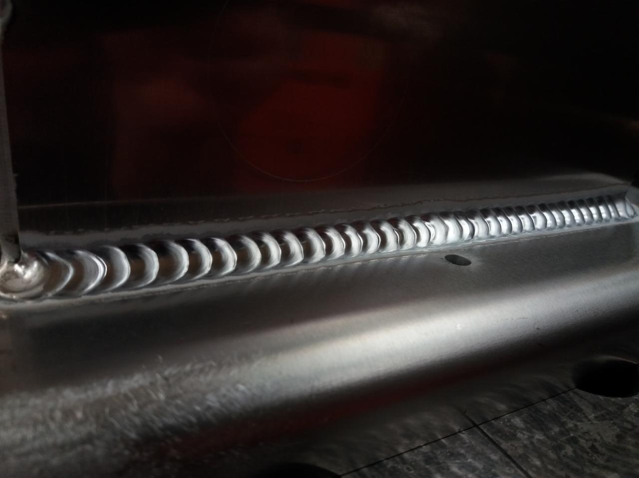 Błyszcząca, aluminiowa powierzchnia z precyzyjnym, regularnym spoiną w kształcie półksiężyców, widoczny otwór w metalu, ciemne tło.