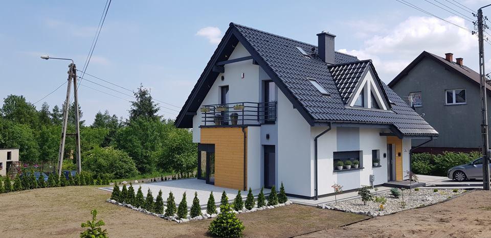 Nowoczesny dom jednorodzinny z balkonem, wykończony w bieli i szarości z elementami drewna, czarny dach i okna, zadbany trawnik i nasadzenia, widok z zewnątrz w słoneczny dzień.