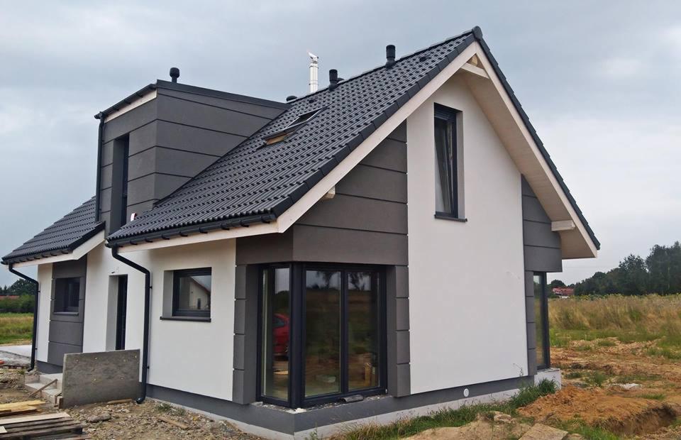 Nowoczesny dom jednorodzinny z ciemną dachówką i elewacją w odcieniach szarości i bieli, widoczny z zewnątrz, z niedokończonym otoczeniem.
