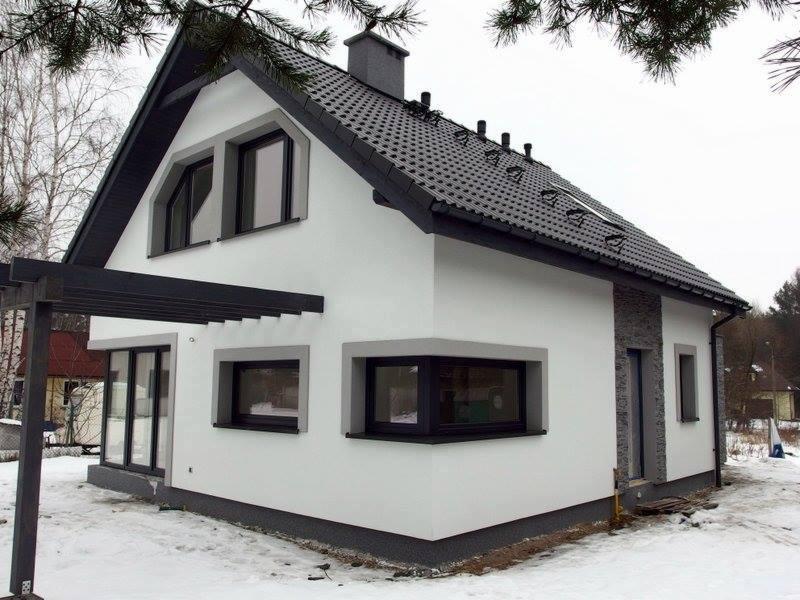 Nowoczesny, dwukondygnacyjny dom z białym tynkiem, ciemną dachówką i grafitowymi ramami okiennymi, z elementami dekoracyjnymi z kamienia na elewacji, widziany w zimowej scenerii.