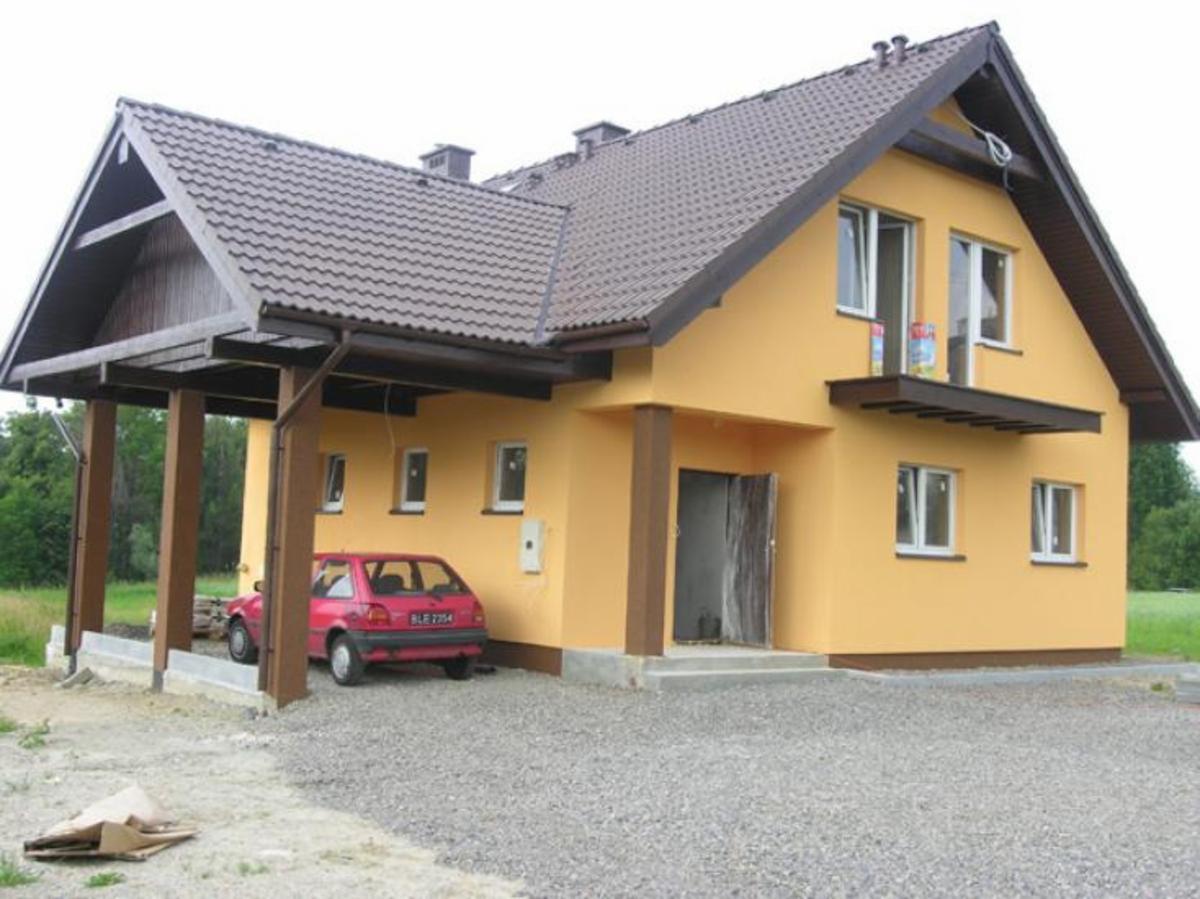 Żółty dom murowany z brązowym dachem i wiatą garażową z czerwonym samochodem, widoczne niedociągnięcia w wykończeniu elewacji.