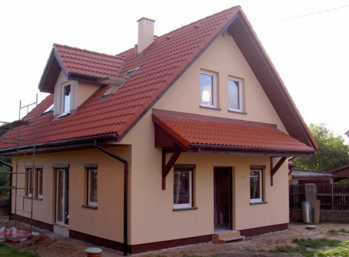 Dwukondygnacyjny dom jednorodzinny z czerwonym dachem, beżową elewacją i widocznym rusztowaniem z boku, sugerującym prace wykończeniowe.