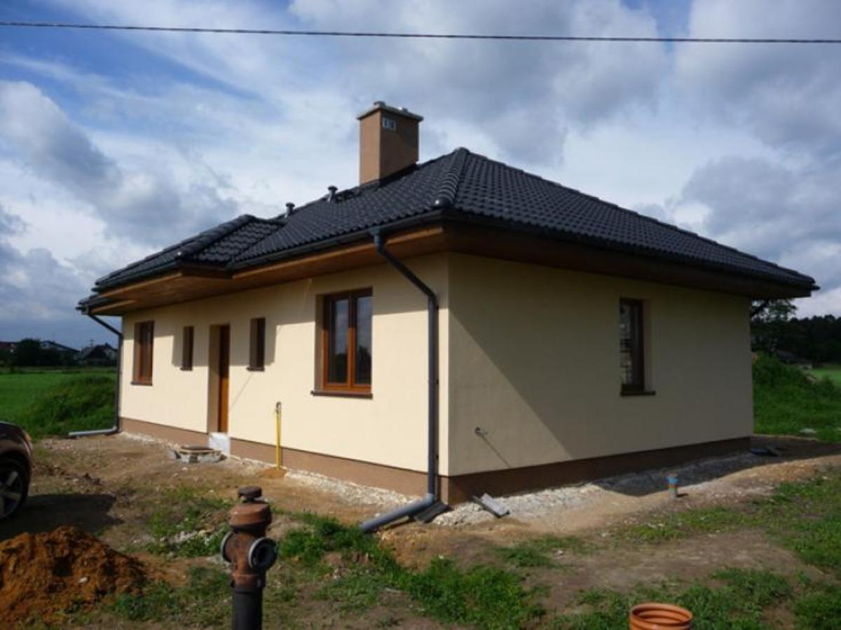 Niedawno wybudowany dom jednorodzinny z ciemną dachówką i beżową elewacją, widoczny z boku, z fragmentem samochodu po lewej stronie i hydrantem na pierwszym planie.