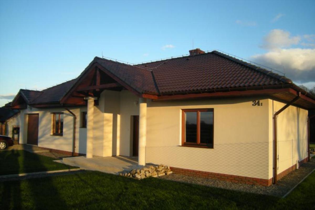 Parterowy dom z beżową elewacją, brązowym dachem i stolarką okienną, widoczne fragmenty muru z cegły klinkierowej przy gruncie oraz numer domu '34₁'.