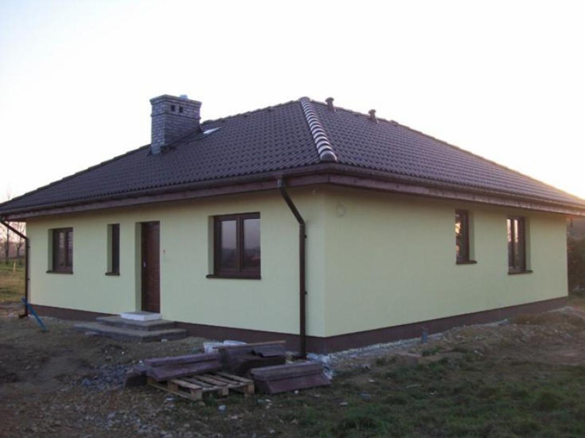 Gotowy dom jednorodzinny z ciemnobrązową dachówką i kominem z cegły, jasna elewacja, widoczne pozostałości materiałów budowlanych na działce.