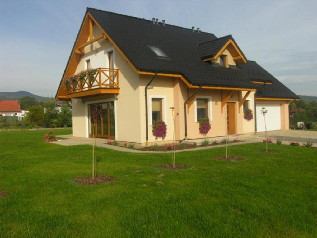 Gotowy dom jednorodzinny z balkonem ozdobionym kwiatami, czarnym dachem i beżową elewacją, otoczony trawnikiem z młodymi drzewkami.