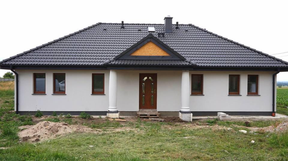 Parterowy dom z ciemną dachówką, drewnianymi oknami i kolumnami przy wejściu, widoczny stos kostki brukowej i niedokończone otoczenie.