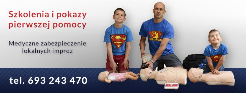 Instruktor demonstruje resuscytację krążeniowo-oddechową na fantomie, obok dwóch chłopców w koszulkach z logo Supermana, imitujących ćwiczenie na innych fantomach.