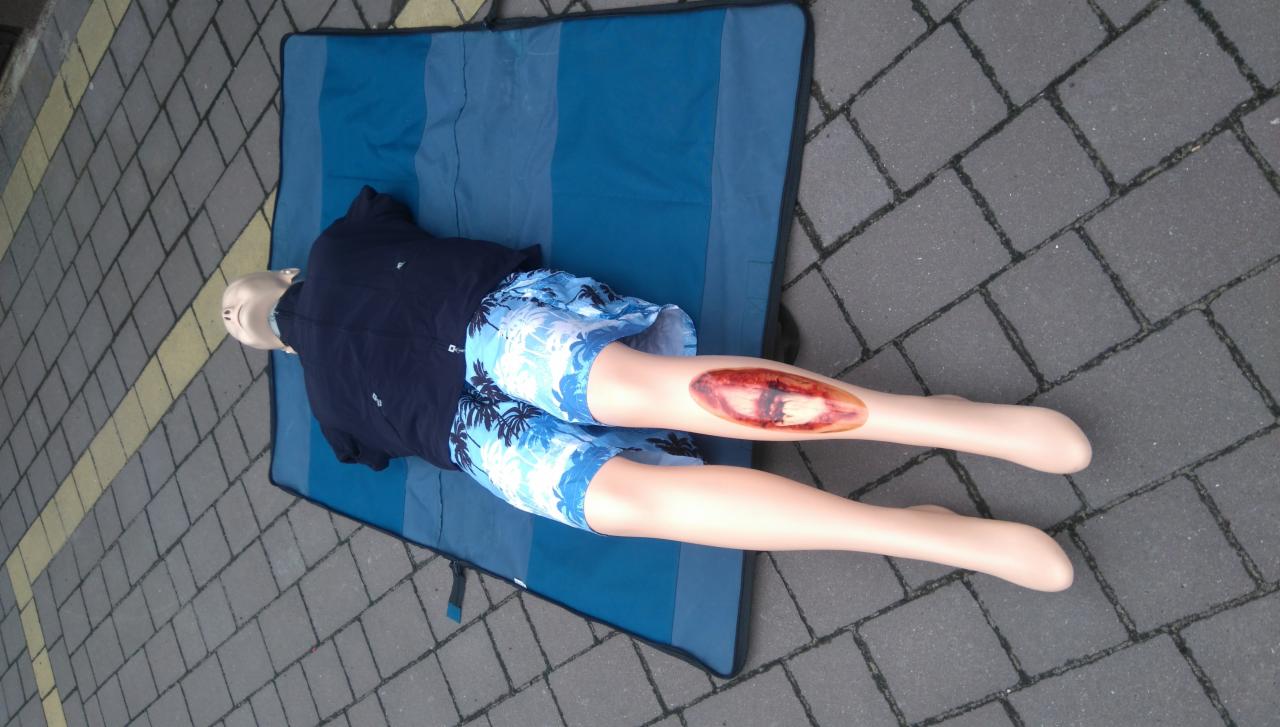 Manekin z symulacją otwartej rany na podudziu, leżący na niebieskiej macie na kostce brukowej.