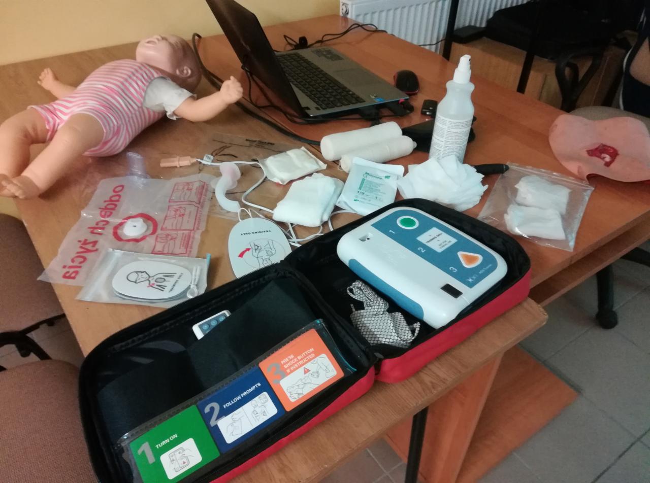 Manekin niemowlęcia leżący obok otwartej walizki z defibrylatorem AED i instrukcjami, bandaże, gaziki, maseczka do resuscytacji, laptop na drewnianym stole.