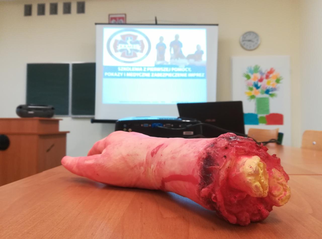 Manekin odciętej ręki na stole podczas szkolenia z pierwszej pomocy, na tle projektora z prezentacją i sali lekcyjnej.